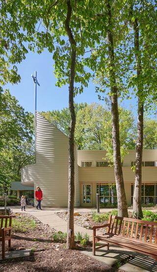 A modern, church building nestled among trees, with two benches in view and a person interacting with a child nearby.