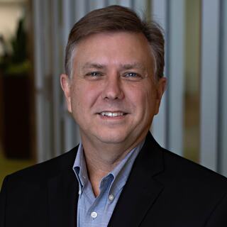 A smiling man with short hair wearing a black blazer over a collared shirt stands in an office setting with blurred background elements.