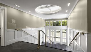A well-lit staircase descends into a spacious hallway, featuring elegant white paneling and large windows with a circular skylight above.