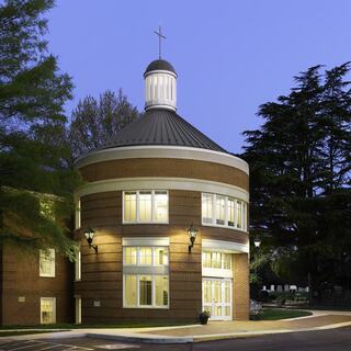 A circular brick rotunda with a domed roof and a cross on top, surrounded by greenery and illuminated by soft lights at dusk.