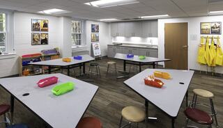 A church classroom features colorful storage bins, tables, and stools, with educational posters on the walls and vibrant aprons hanging nearby.