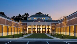A modern church building featuring brick, large glass doors, surrounded by landscaped greenery and evening twilight, creating a welcoming ambiance.