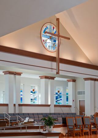 A bright sanctuary with high ceilings and elegant wood accents, featuring a large cross and colorful stained glass windows.