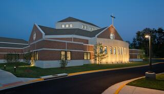 Heritage Fellowship church exterior features brick and stone architecture, illuminated at dusk, with a cross atop and landscaped surroundings.