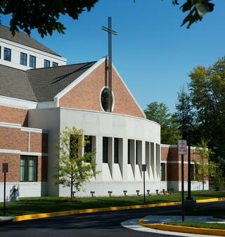 A modern church exterior features a large cross, brick and white facade, surrounded by greenery and a clear blue sky, creating a tranquil atmosphere.