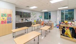 A bright and cheerful classroom featuring tables, artwork on the walls, a kitchenette area, and colorful educational materials.