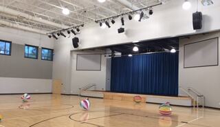A spacious multipurpose space/gymnasium features a stage with blue curtains, bright lights, and colorful beach balls scattered on the polished wooden floor.