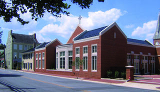 A historic brick church with large windows stands beside an older green historic structure under a blue sky with scattered clouds.