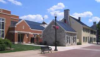 A mix of modern and traditional architecture lines a sunny street, featuring greenery, a bench, and colorful flower baskets.
