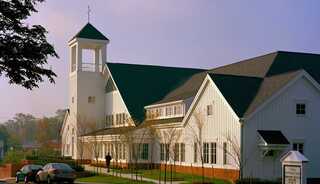 A white church building features a tall bell tower and a sloped roof, surrounded by trees and a walkway. Soft morning light enhances its charm.