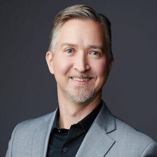 A smiling man with gray hair and a well-groomed beard wears a light gray suit over a black shirt, showcasing confidence and approachability.