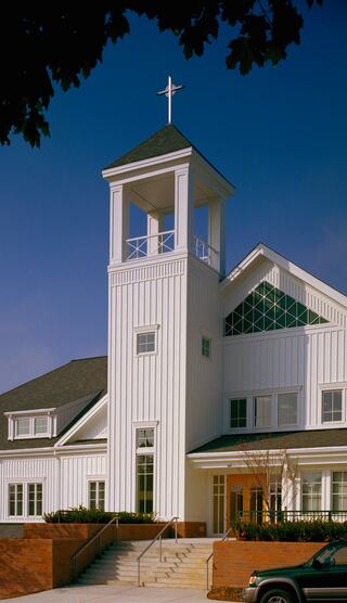 A white church with a tall steeple and a cross, featuring large windows and a welcoming entrance surrounded by greenery.
