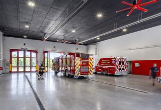 A spacious fire station interior with red and white fire trucks, large doors, and a clean, modern design. Ceiling fans enhance airflow.