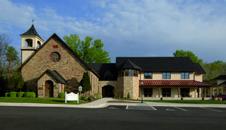 A charming stone church with a bell tower is complemented by a modern adjacent building, set against a backdrop of green trees and blue skies.