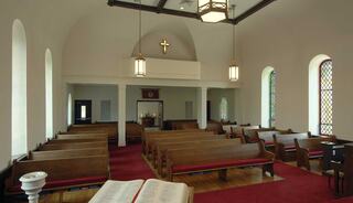 A church interior featuring wooden pews, stained glass windows, red carpeting, and a central altar with soft lighting.
