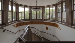 A wooden staircase with railings and large windows, creating a bright, open atmosphere, overlooks lush greenery outside.