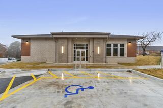 A modern building with a light-colored stone facade, large windows, and accessible parking, set against a clear sky.