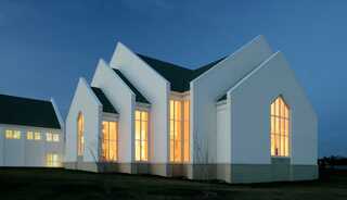 A modern, white church building with angular roofs and warm, glowing windows, set against a twilight sky.