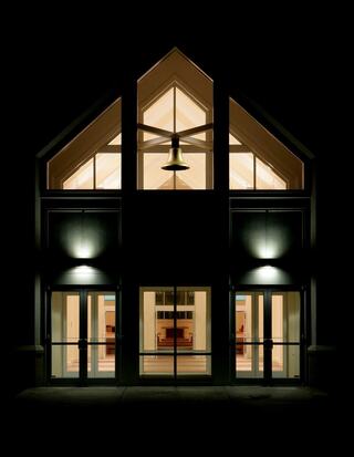 A modern church building features a triangular roof with a prominent bell at the center. Warm light glows from within, highlighting the entrance.