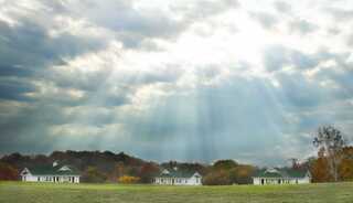 A tranquil landscape features several church cabins on a grassy field, illuminated by soft rays of sunlight breaking through cloudy skies.