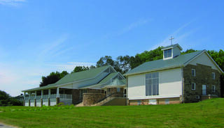 A serene chapel and multipurpose space featuring a renovated stone barn with a glass facade and a traditional structure, set against a clear blue sky and green grass.