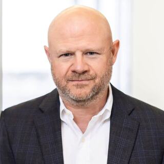 A confident man with a bald head and a beard wears a dark blazer over a white shirt, standing against a bright, blurred background.