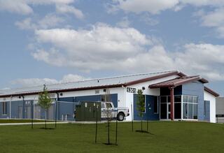 A modern, low-rise building with a blue exterior, red roof, and large windows, surrounded by grassy landscaping and small trees.