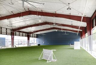 A spacious indoor facility featuring artificial turf, high ceilings, large fans, and open areas, ideal for sports or activities.