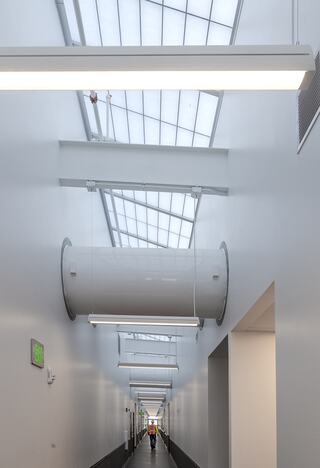 A long, modern hallway features large skylights and overhead lighting, leading to a distant figure, with sleek white walls and minimal design.