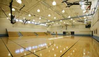 A spacious gymnasium features polished wooden floors, bright lighting, and bleachers along the walls, ready for sporting events or activities.