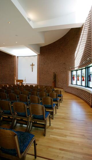 A serene chapel featuring wooden floors, wooden chairs, artistic stained glass windows, and a prominent cross on the wall.