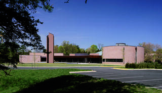 A modern chuch building features unique architectural shapes and a mix of brick and glass, set against a clear blue sky and green landscaping.