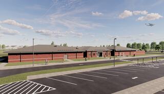 A modern, single-story building features brick architecture, surrounded by trees and a parking lot, with an airplane flying overhead.