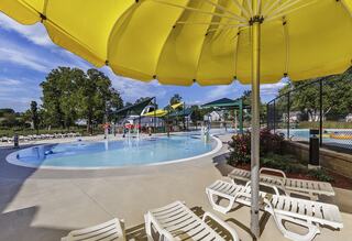 A vibrant pool area features a large yellow umbrella, lounge chairs, playful water slides, and lush greenery, creating a fun, inviting atmosphere.