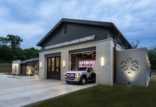 A modern ambulance station with a large garage, featuring a lit ambulance and sleek architecture, set against a twilight sky.