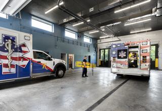 A spacious emergency vehicle bay features two ambulances, one being loaded with equipment, and a worker preparing for a call.