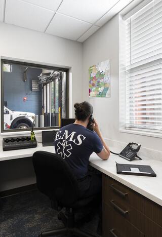 An EMS worker sits at a desk in an office, engaged on the phone, with maps and emergency vehicles visible through a window.