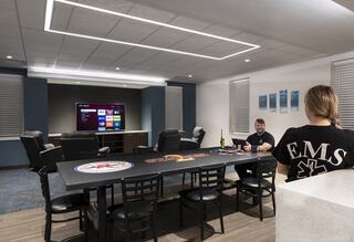 A modern lounge features a large table, seating, and a wall-mounted TV, with two people engaged in conversation and a relaxed atmosphere.