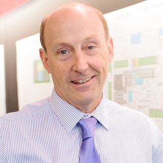 A man smiles confidently, wearing a striped shirt and a tie. He stands in front of a backdrop featuring various colorful plans or diagrams.
