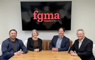 Four professionals are seated at a table, smiling, with a large screen behind displaying "fgma FGMA Architects."