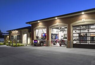 A modern emergency service building at dusk, featuring illuminated garage doors with emergency vehicles parked outside.