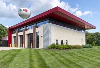 A modern building with a red roof and large windows, surrounded by well-maintained grass and trees, featuring a water tower nearby.