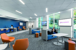 A modern, well-lit lounge features comfortable seating, desks, and two large screens. Large windows provide natural light and a welcoming atmosphere.