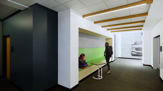 A modern office interior features a colorful seating area, wooden beams on the ceiling, and a person sitting while another stands nearby.