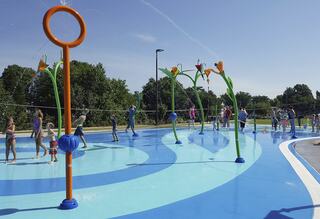 Children play joyfully in a vibrant splash pad, surrounded by colorful water features and a sunny, clear blue sky.