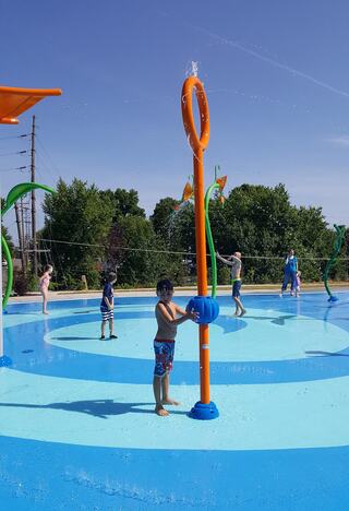 Children play in a colorful splash pad, some interacting with water features under a clear blue sky, surrounded by greenery.