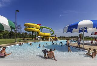 A lively pool scene with children and families enjoying water activities, featuring colorful slides and shaded umbrella areas under a clear sky.