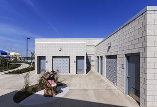 A sunny outdoor area features a block structure with lockers and a playful statue, surrounded by neatly landscaped pathways and vibrant colors.