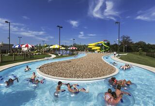 A vibrant water park scene featuring a lazy river with floating visitors, colorful umbrellas, and an exciting yellow waterslide under a clear blue sky.