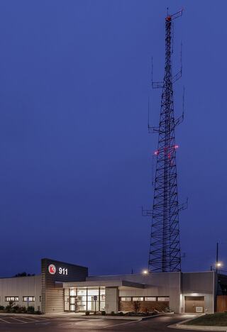 A modern emergency services building with a tall communication tower, illuminated under a blue twilight sky. The "911" sign is prominent.